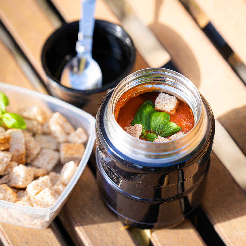 Een geopende thermoskan toont verse tomatensoep met basilicum en croutons. Naast de thermos staat een bakje met extra croutons en een lepel.