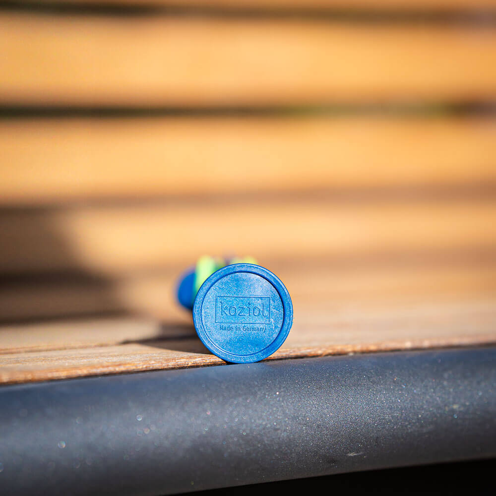 Close-up van de onderkant van een blauw stuk bestek dat op een houten bankje ligt. De onderkant toont het logo "Koziol Made in Germany."