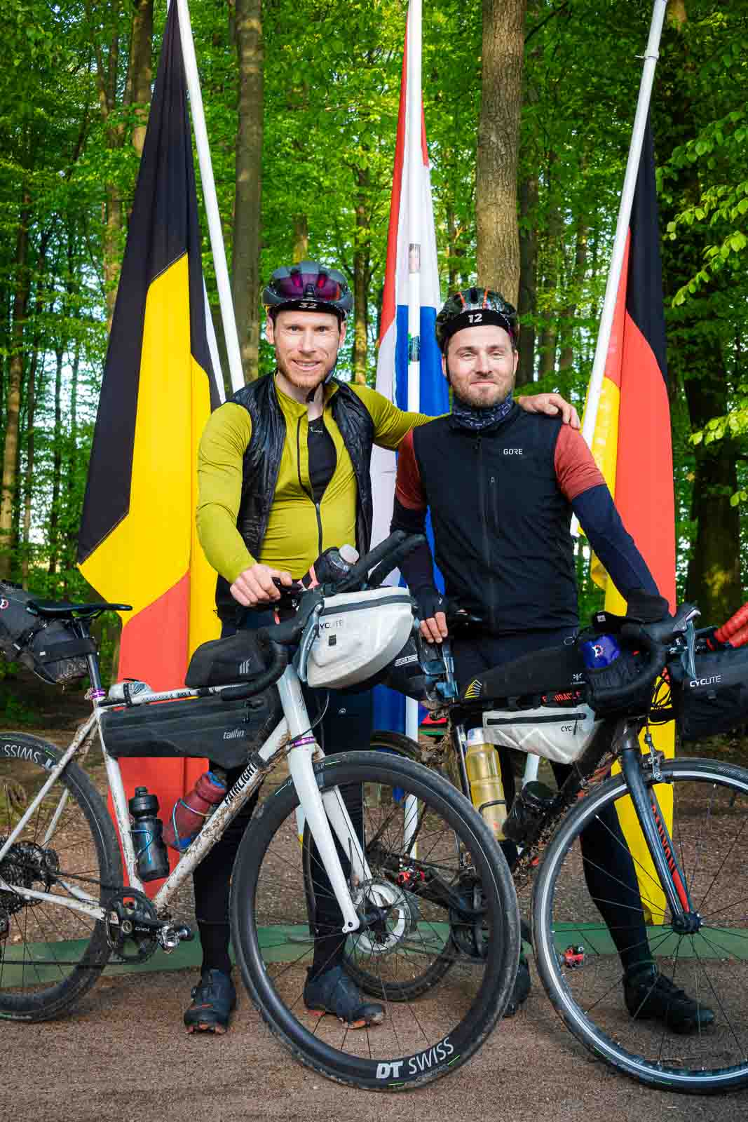 Twee fietsers staan bij het drielandenpunt van Duitsland, België en Nederland en lachen in de camera.