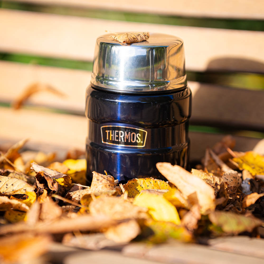 Een blauwe thermoskan staat op een houten bank, omringd door herfstbladeren. Op het glanzende deksel ligt een klein blaadje. De kan draagt het logo "Thermos" en schittert in de herfstomgeving.