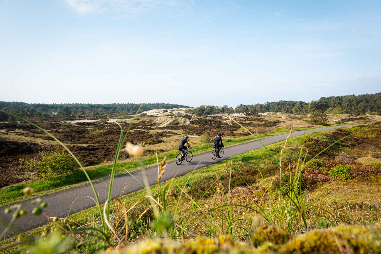 Twee fietsers die meedoen aan de Race om Nederland rijden over een weg door de duinen.