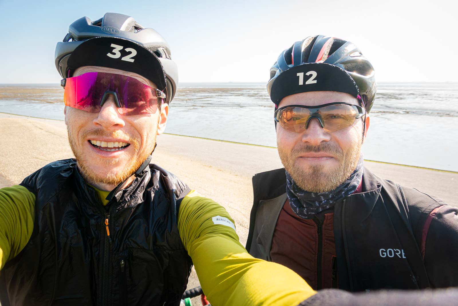 Twee fietsers nemen een selfie van zichzelf terwijl ze voor een strandpanorama staan en lachen.